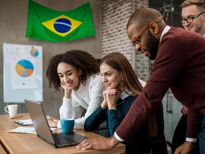 grupo de colegas en una oficina colaborando frente a una laptop. Se les ve sonrientes y enfocados en la pantalla, mientras que en la pared del fondo destaca una bandera de Brasil