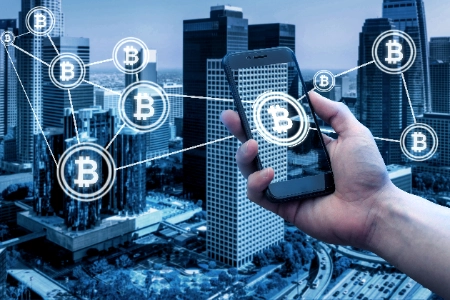 mano sosteniendo un smartphone frente a un paisaje urbano moderno. En la pantalla del dispositivo y flotando sobre los edificios, se observan iconos de Bitcoin interconectados por líneas de red
