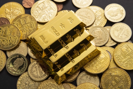 Uma pequena pilha de lingotes de ouro gravados repousa sobre um grande número de moedas de ouro de várias denominações e origens, incluindo uma moeda Bitcoin.