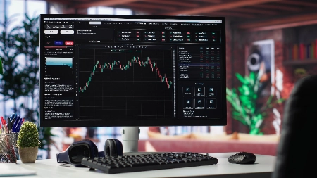 Um monitor de computador em uma mesa de escritório exibe uma plataforma de negociação profissional com gráficos detalhados de velas financeiras, indicadores de mercado e listas de ativos.
