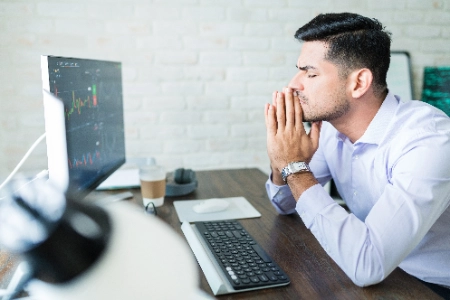 Um homem com expressão de profunda concentração ou estresse observa gráficos financeiros em seu monitor, com as mãos juntas em frente ao rosto enquanto analisa os movimentos do mercado em sua mesa.