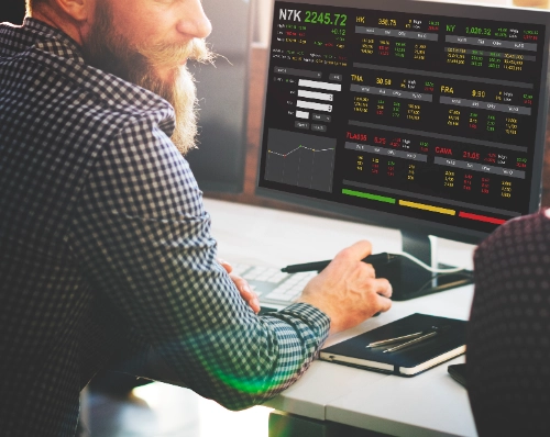 Un hombre de perfil con barba y camisa a cuadros, observando una pantalla de computadora que muestra un panel de control financiero con datos de bolsa de valores, gráficos e indicadores de mercado.