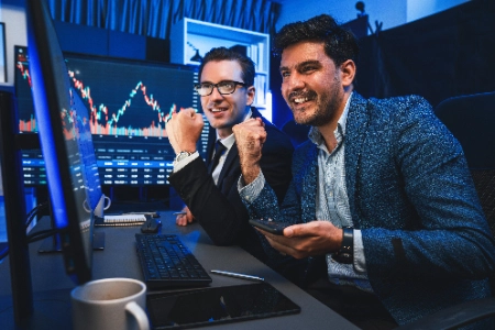dos personas colaborando en el análisis de inversiones. Mientras una sostiene un smartphone que muestra un gráfico de trading, la otra utiliza una laptop para comparar datos en tiempo real