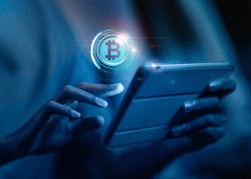 Primeiro plano em tons azulados de mãos a segurar e a tocar num tablet, do qual emerge um holograma brilhante com o logótipo da Bitcoin.