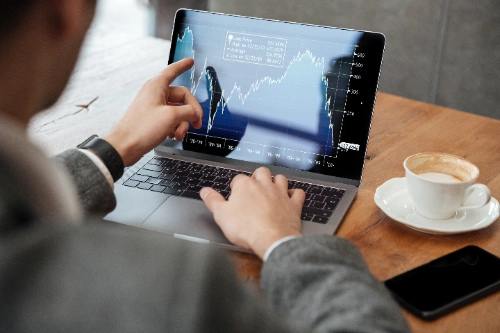 hombre con chaqueta gris sentado a una mesa de madera. Con el dedo índice de su mano izquierda señala un punto específico en un gráfico financiero detallado que se muestra en la pantalla de su laptop