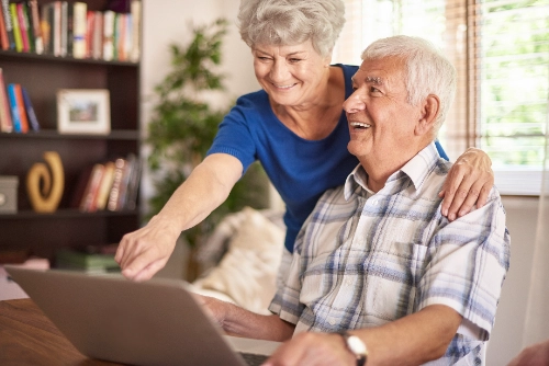 se muestra a una pareja de adultos mayores sonriendo alegremente frente a una computadora portátil en lo que parece ser una sala de estar.
