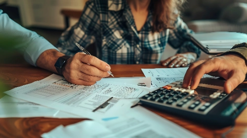 se muestran las manos de dos personas trabajando en un escritorio de madera: una mano sostiene un bolígrafo sobre unos documentos impresos, mientras que otra mano utiliza una calculadora en primer plano a la derecha.