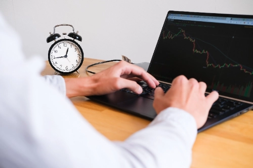 Primer plano lateral de las manos de un hombre escribiendo en una computadora portátil que muestra un gráfico financiero descendente, con un reloj despertador clásico negro sobre la mesa