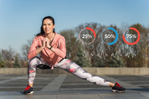 Una mujer al aire libre realiza un estiramiento lateral de piernas en ropa deportiva, acompañada de gráficos digitales flotantes que muestran indicadores de ritmo cardíaco y porcentajes