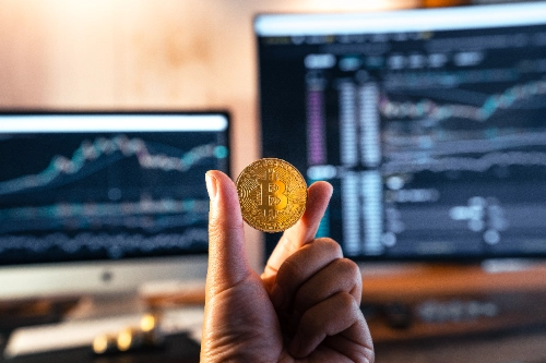 Una mano sostiene una moneda física de Bitcoin en primer plano, con dos pantallas de ordenador de fondo mostrando gráficos de trading difuminados.