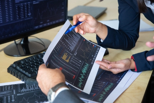 Dos analistas financieros revisan y señalan reportes impresos con gráficos bursátiles en un escritorio de trabajo, junto a pantallas digitales con datos de mercado.