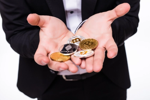 Un hombre en traje sostiene en sus manos abiertas varias monedas físicas de criptomonedas, incluyendo Bitcoin y Ethereum.