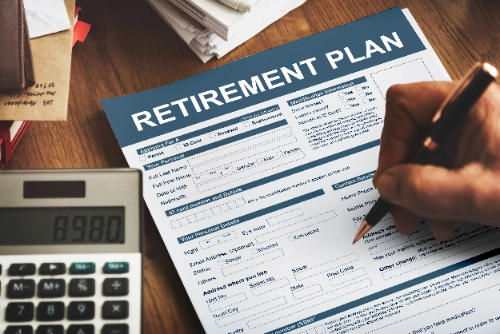 Una mano escribe con un bolígrafo sobre un formulario titulado "RETIREMENT PLAN". En la esquina inferior izquierda se observa una calculadora digital.