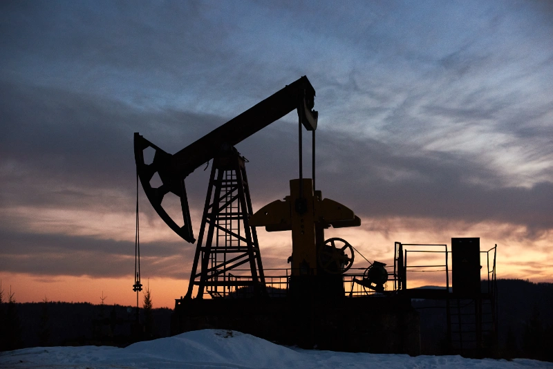 La silueta de una bomba de varilla (balancín de extracción petrolera) se recorta contra el cielo al anochecer, con tonos anaranjados y azulados en el horizonte y nieve en el suelo.