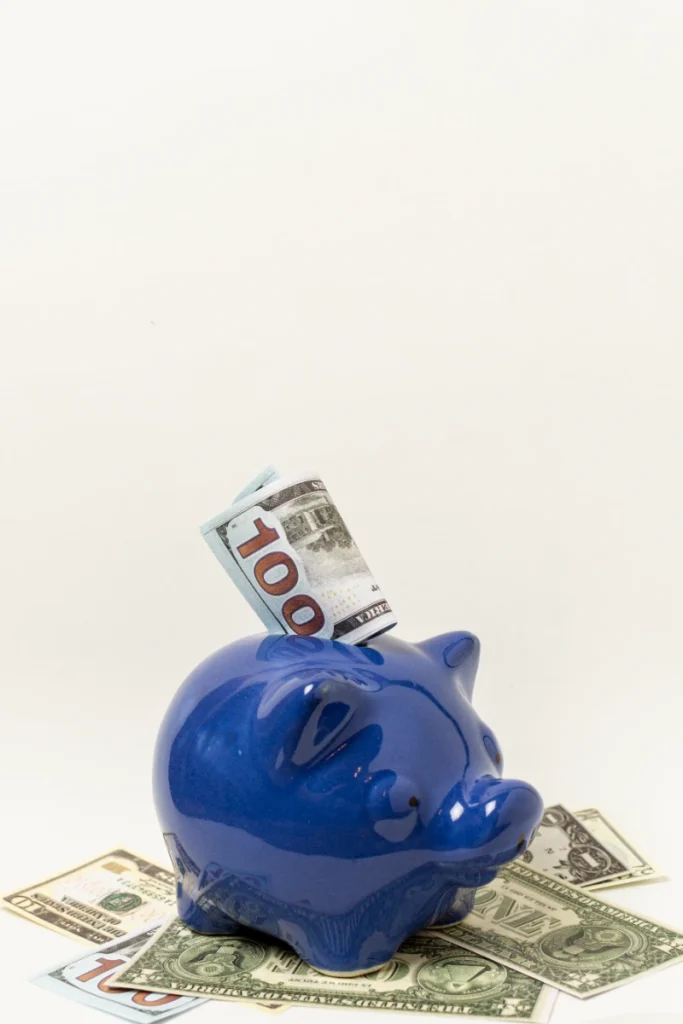 Un alcancía de cerdito azul con un billete de 100 dólares en la ranura, rodeada de dinero sobre un fondo claro.
