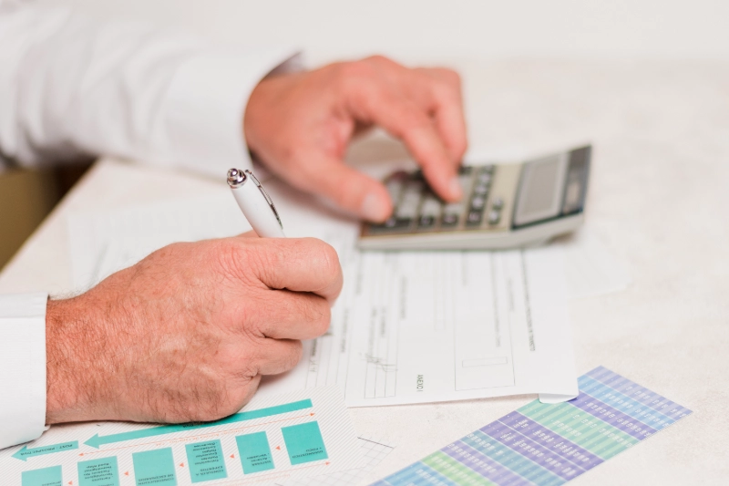 Manos de un profesional escribiendo con un bolígrafo en documentos financieros mientras utiliza una calculadora sobre el escritorio.