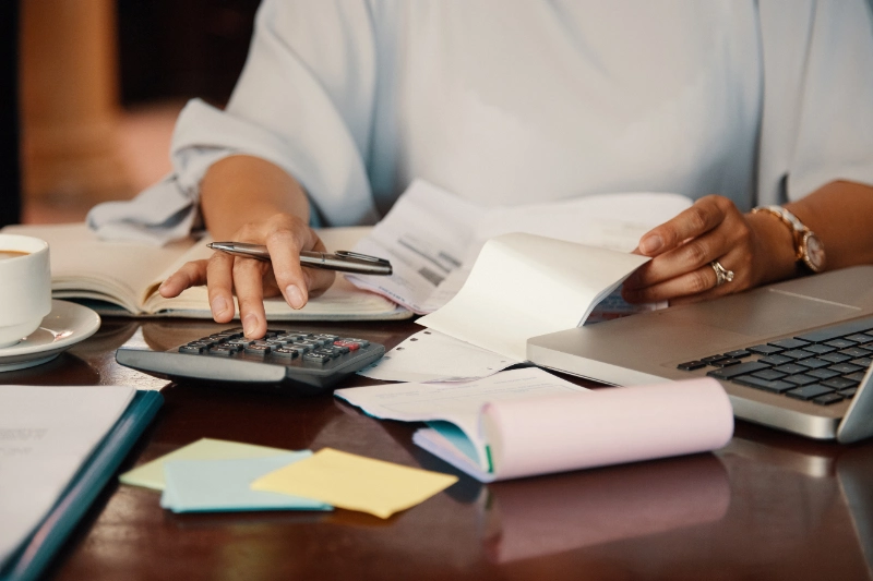 Persona utilizando una calculadora y revisando facturas e impresos en un escritorio junto a una laptop y una taza de café.